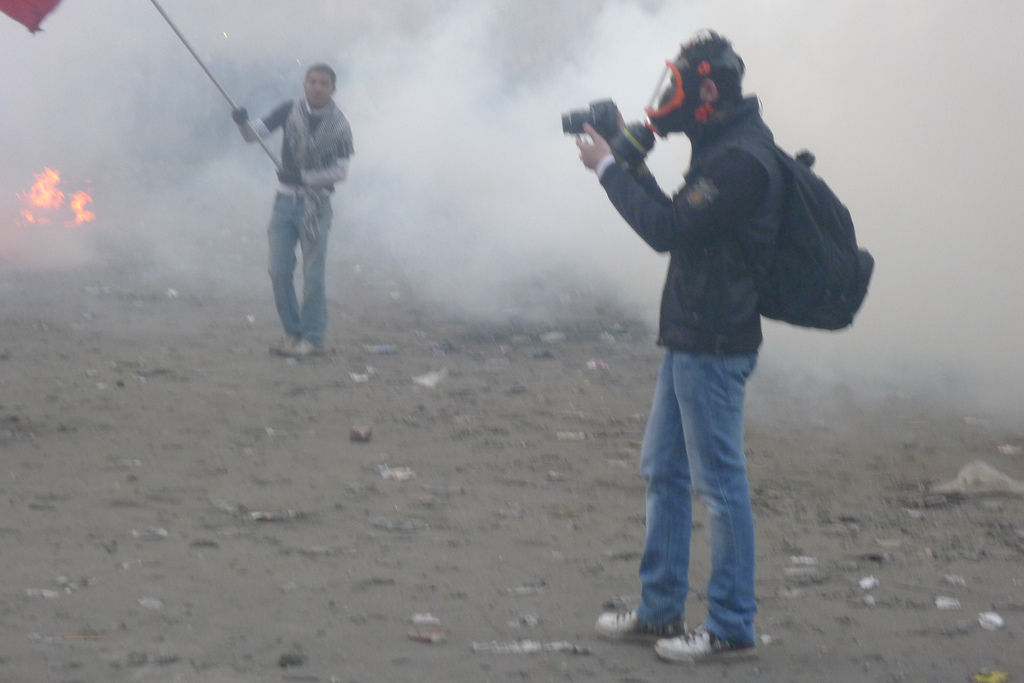 Person wearing gas mask and taking photos, surrounded by clouds of gas, with flames and flag-waving demonstrators in the background