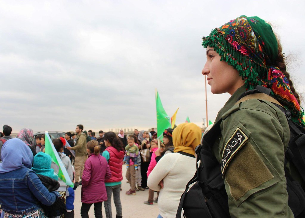 A woman in military fatigues and traditional headscarf overlooks a group of civilians of all ages.