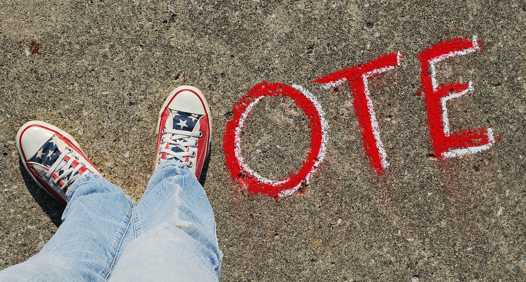 Feet in red, white and blue tennis shoes form a letter V, followed by the letters O-T-E in chalk on the sidewalk