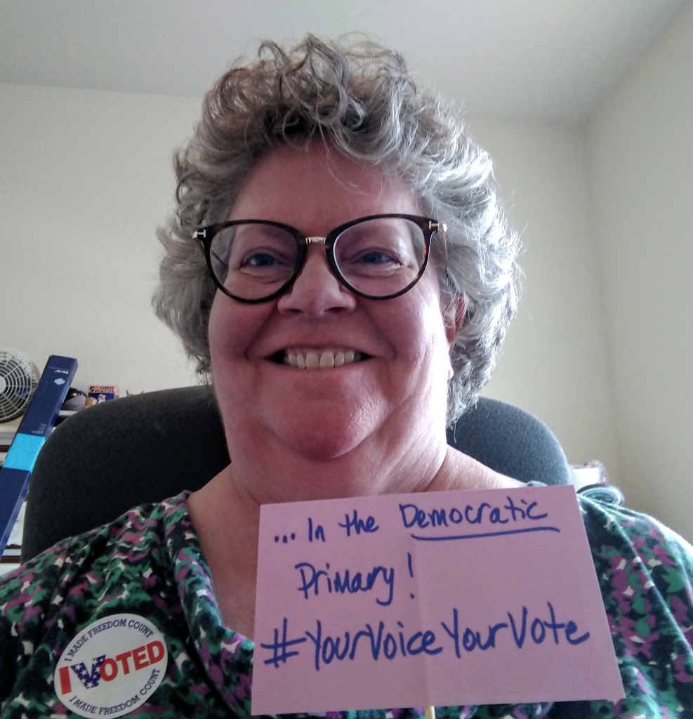 Laura Bray smiles as she wears her "I Voted" sticker, holding a sign that reads "...in the _Democratic_ primary! #YourVoiceYourVote"