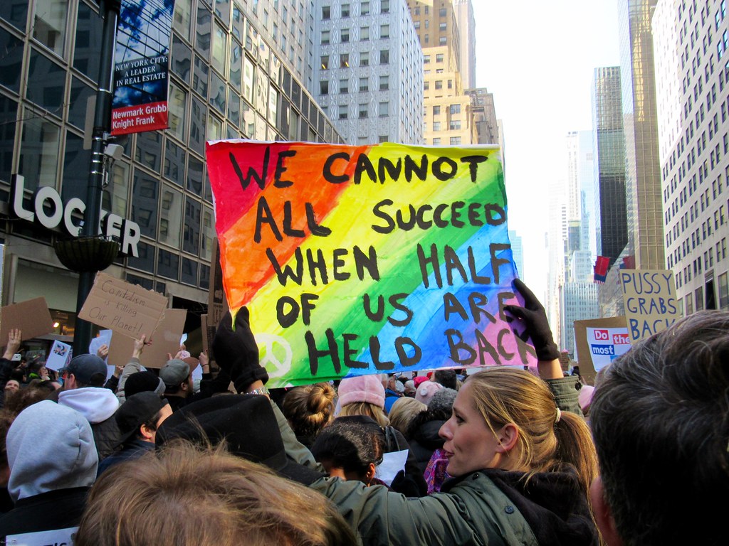 In the midst of a crowd, a woman holds up a rainbow-colored sign reading "We cannot all succeed when half of us are held back."