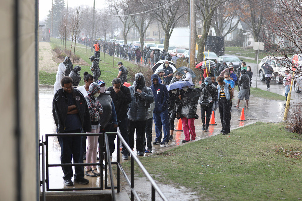 A long line of voters, many wearing face coverings, stretches up the street in the midst of a rainstorm.