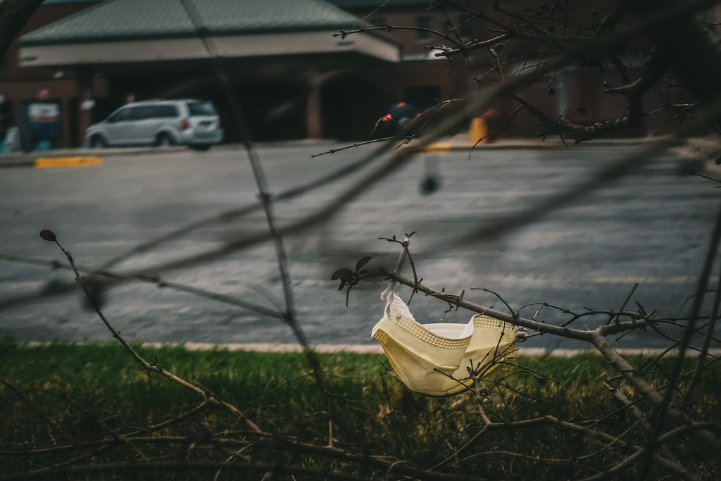 A discarded facemask is tangled in a bush, with parking lots and buildings in the background