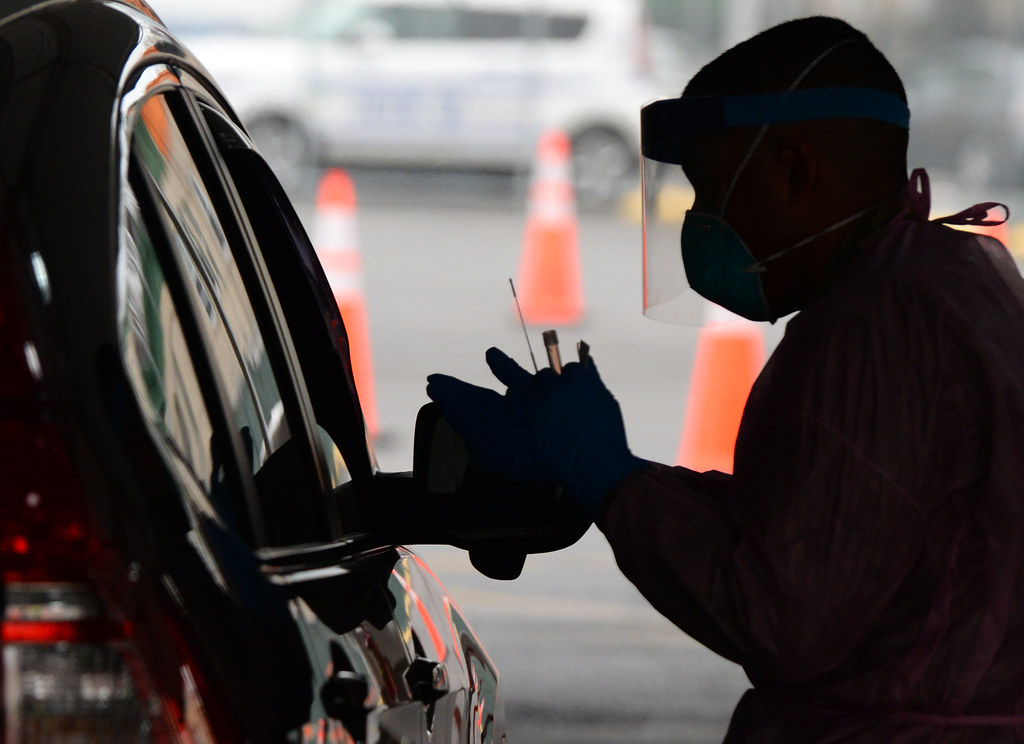 A national guard member in mask and face shield steps up to the passenger window of a car, with testing swab and vial in hand.