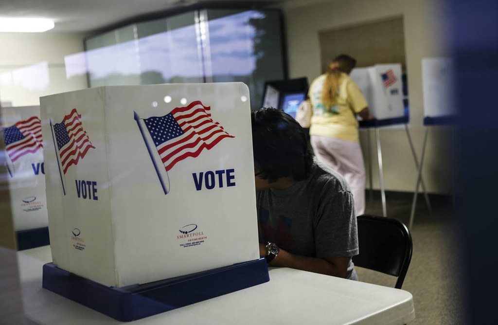 A voter sits at a voting enclosure while another stands at a voting machine in the distant background