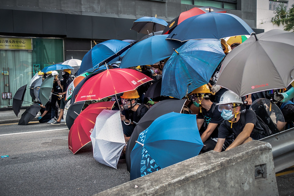 Protestors with masks and hardhats form a line, protecting themselves with umbrellas