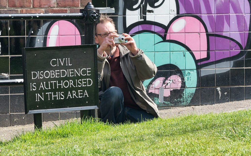 Man taking a picture in front of a sign which reads "Civil disobedience is authorised in this area"