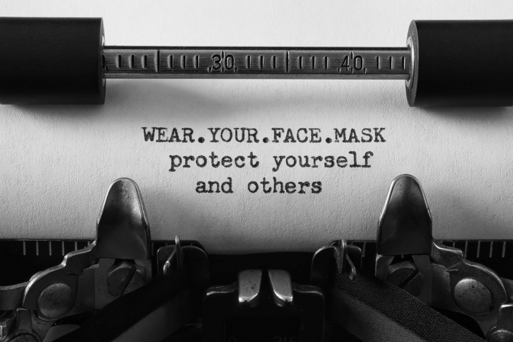 Closeup of a typewritter and paper with the message "WEAR.YOUR.FACE.MASK protect yourself and others" typed on the paper.