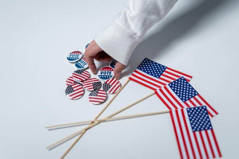 Small American flags and small flag buttons and Vote buttons on surface with hand reaching to pick up one Vote button.